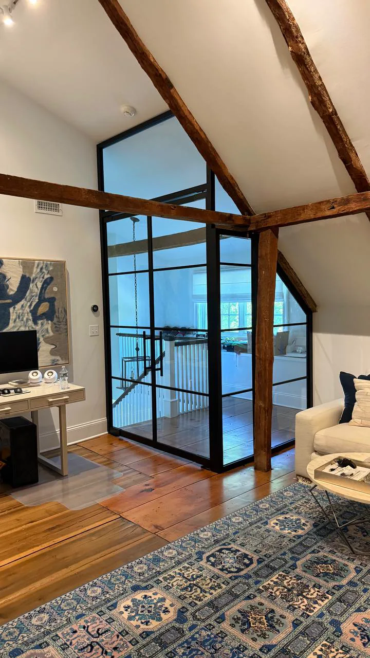 Glass partition raked to a barn roof pitch with French doors, threaded past hand-hewn tie beams in a Connecticut loft by WiseGlass NY