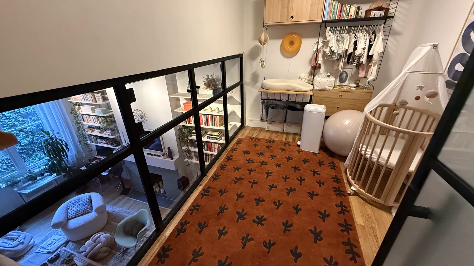 Upper-floor room behind a glass wall on black structural frames with opening windows, looking down to the living room below, Manhattan duplex by WiseGlass NY